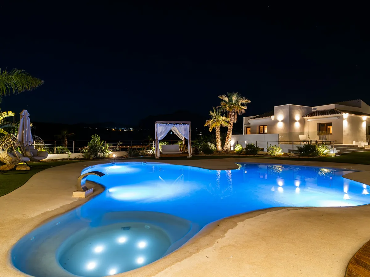 A freeform pool glowing blue at night beside a lit villa